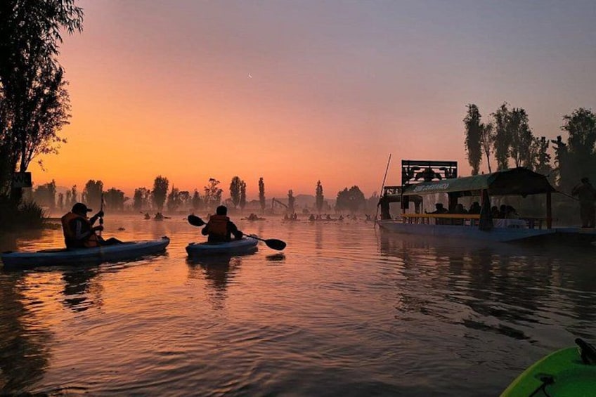 Kayakers on a calm river at sunset, a boat docked on the right, and trees lining the riverbank. 1