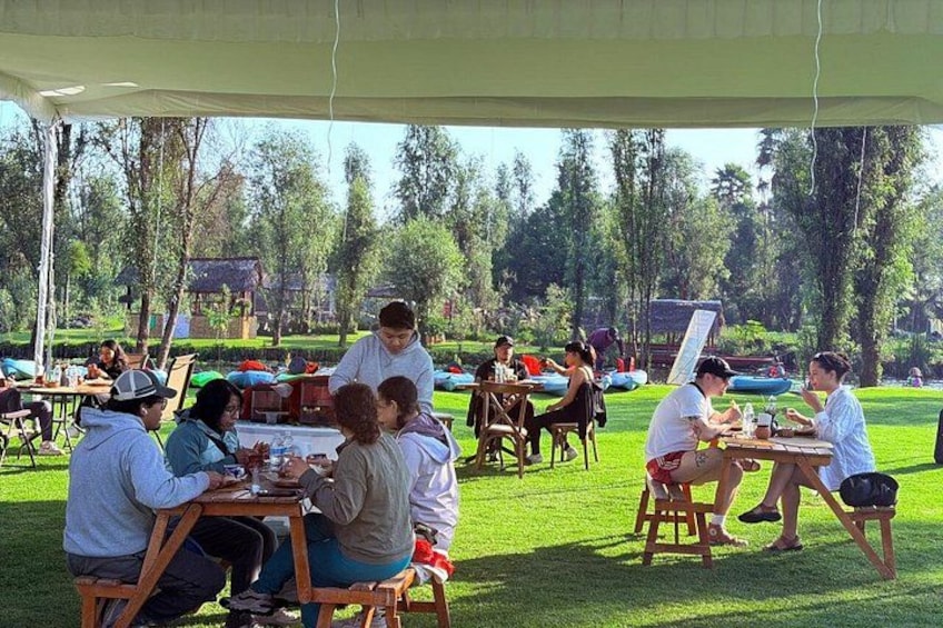 People sitting at tables under a tented area in a park with grass and trees in the background. 4