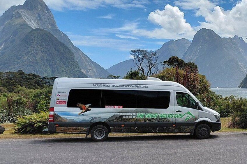 A white bus with 'The Great Walks Bus' branding parked on a road with a mountainous landscape in the background. 1
