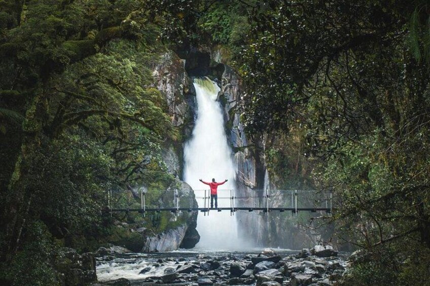 A person standing on a bridge with arms outstretched, overlooking a waterfall surrounded by lush greenery. 3