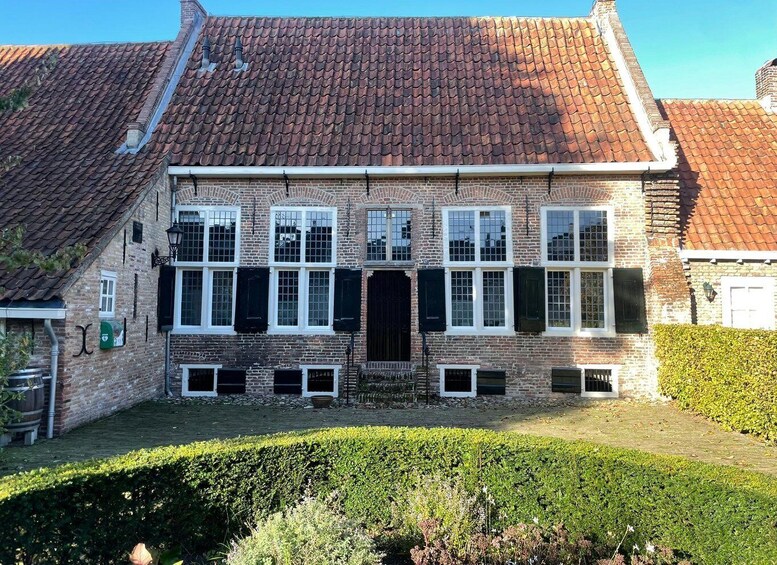 A two-story brick house with a red-tiled roof, white window frames, and a green hedge in the foreground. 1