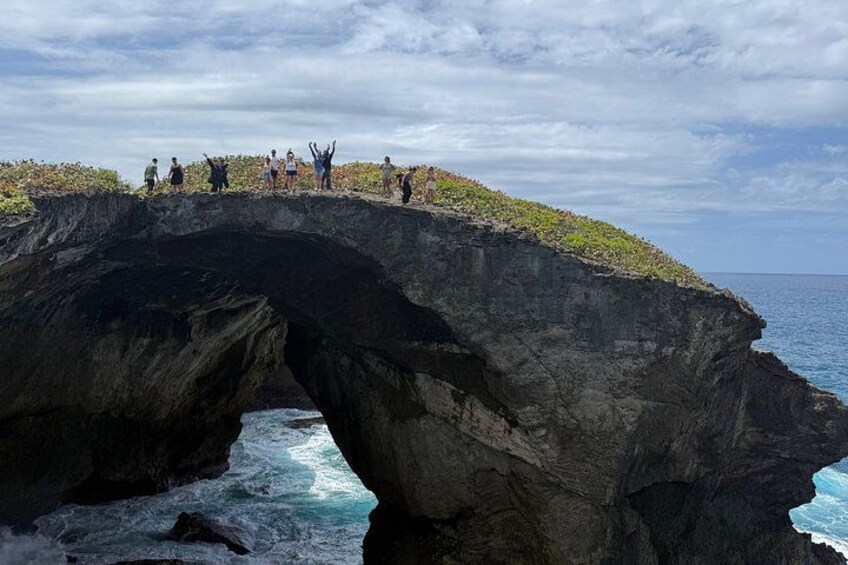 A natural rock formation with a bridge, ocean waves, and people standing on the edge. 1