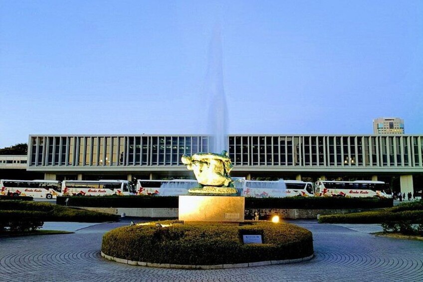 Une fontaine ornée d’une statue au centre, entourée d’un jardin circulaire, avec un bâtiment moderne en arrière-plan. 2