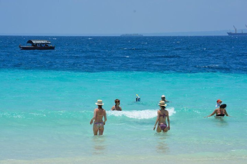 A beach with clear turquoise water, people swimming and walking, and a boat in the distance. 4