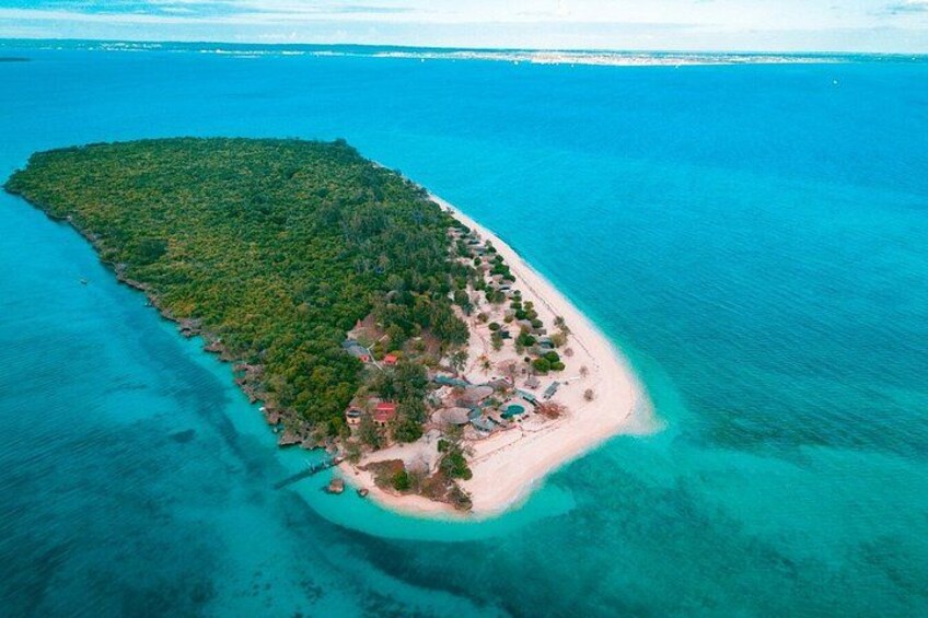 An aerial view of a tropical island with a clear turquoise sea, dense green vegetation, and a small settlement with buildings and a beach. 1