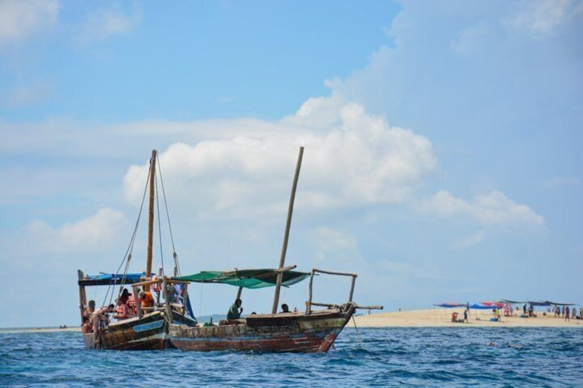 A traditional wooden boat with a sail on the water near a sandy beach with people and umbrellas in the background. 5