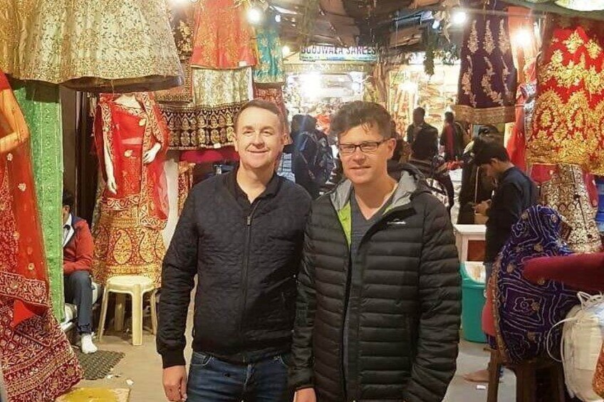 Two people standing in a market with colorful fabrics and textiles on display. 2