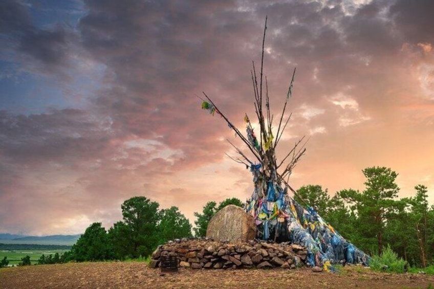 A totem pole with a painted tipi and a stone base, surrounded by trees and a cloudy sky. 3