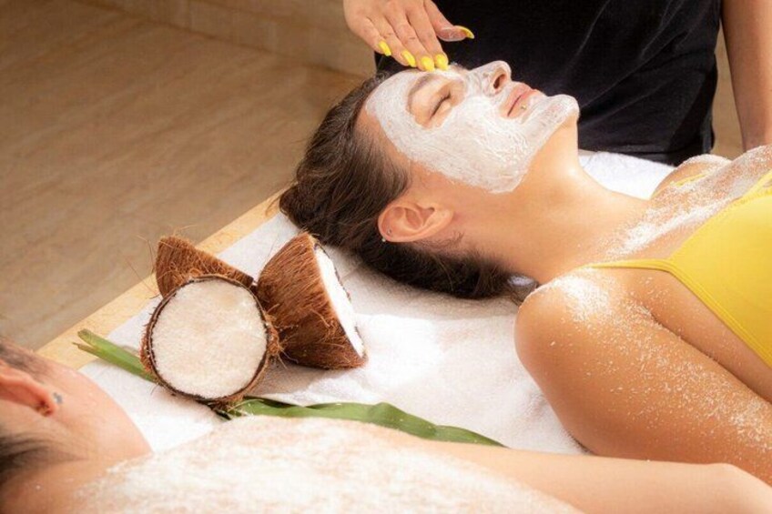 A person receiving a facial treatment with coconut shells and green leaves on a massage table. 4