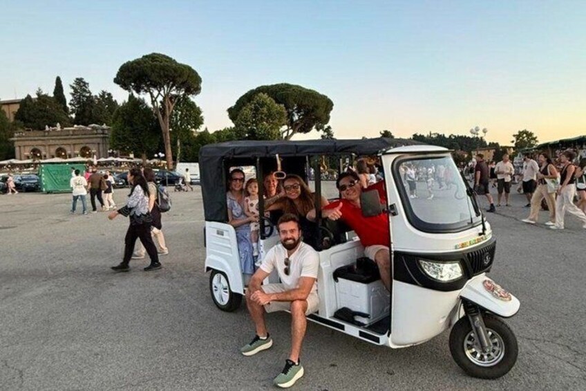 A group of people in a small, open-top vehicle, likely a tuk-tuk, in a crowded outdoor area with trees and buildings in the background. 5