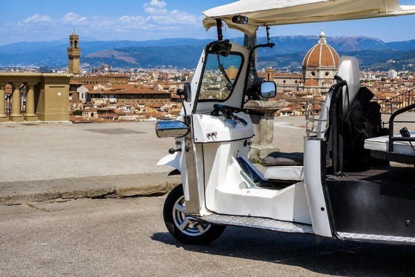 A white three-wheeled vehicle parked on a paved area with a cityscape and a prominent dome-shaped building in the background. 2