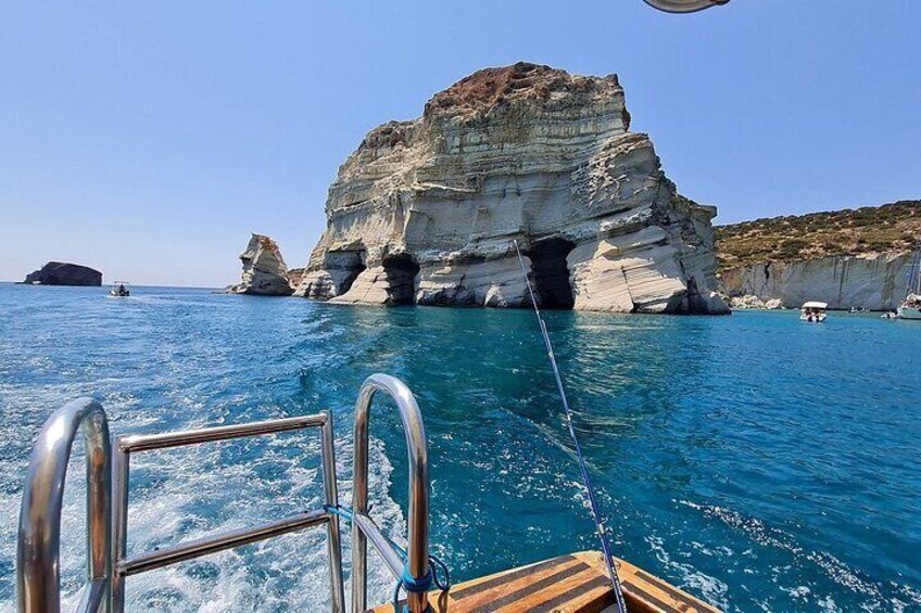 A boat with a metal railing, clear blue sea, and a large rock formation with a cave entrance. 1