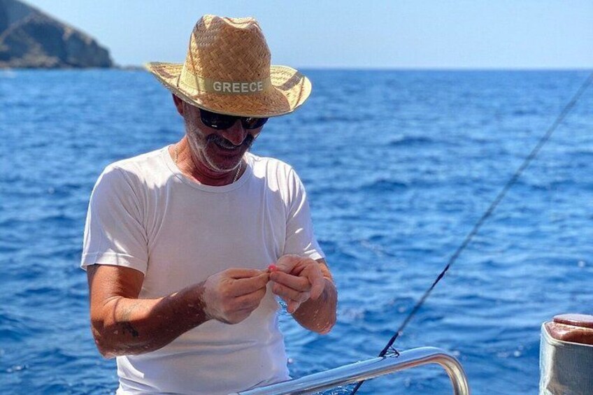 A person on a boat wearing a straw hat with 'GREECE' written on it, holding a fishing rod. 2