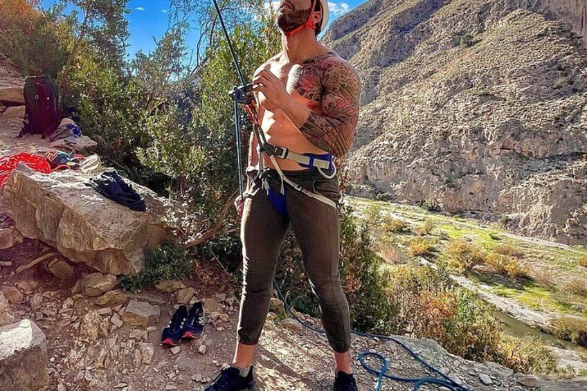 A person with tattoos standing on a rocky terrain, holding a climbing harness and ropes, with a mountainous landscape in the background. 5