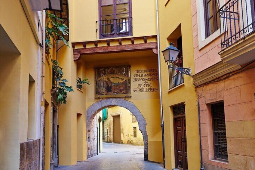 A narrow alleyway with a stone archway, yellow buildings, and a sign above the archway. 4