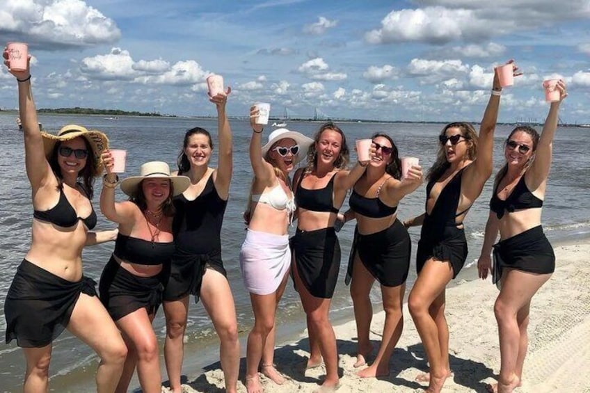 A group of people on a beach, some holding pink cups, with a body of water and a clear sky in the background. 5