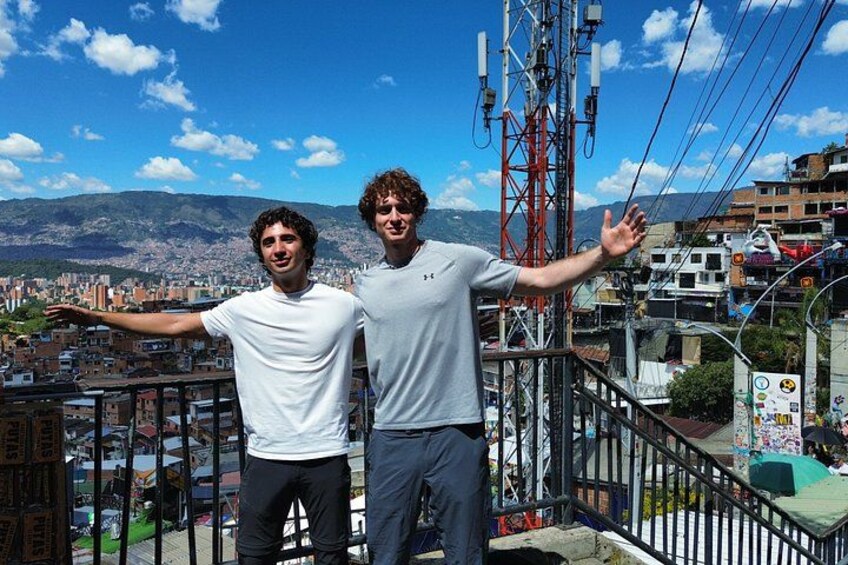 Two people standing on a balcony with a cityscape and mountains in the background. 5
