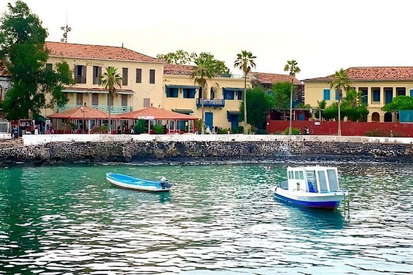 A calm waterfront with two boats, a stone wall, and buildings with red roofs. 1