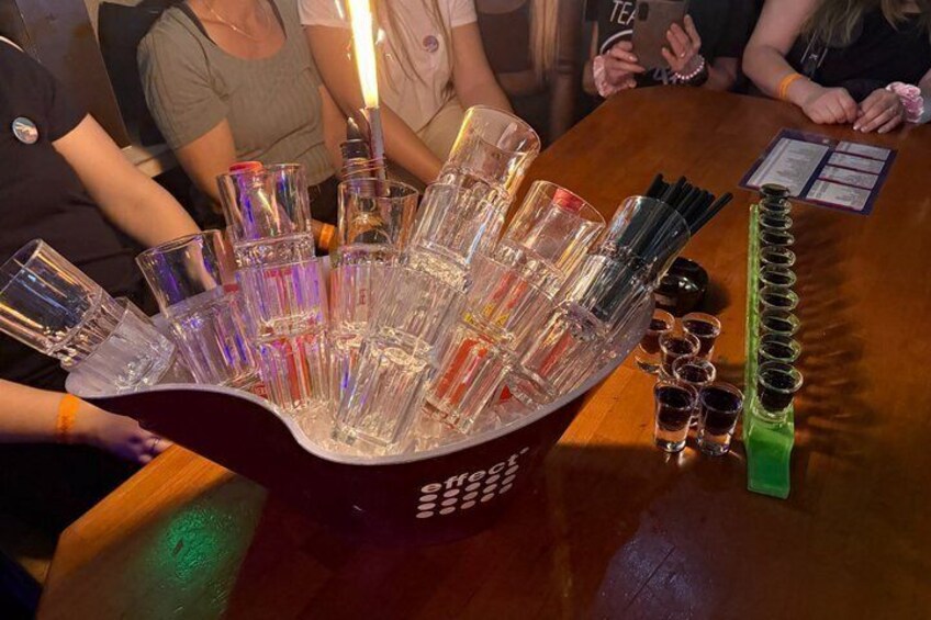 En bar med en stor, genomskinlig glasbehållare fylld med små glas för snaps och ett grönt ställ för att hålla glasen. 5