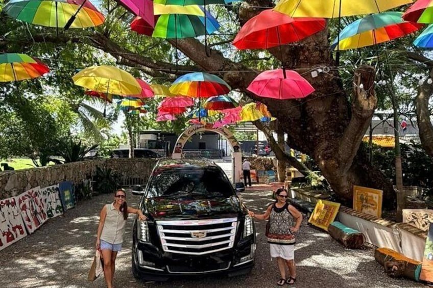A black car parked under colorful umbrellas, surrounded by trees and a stone wall. 5