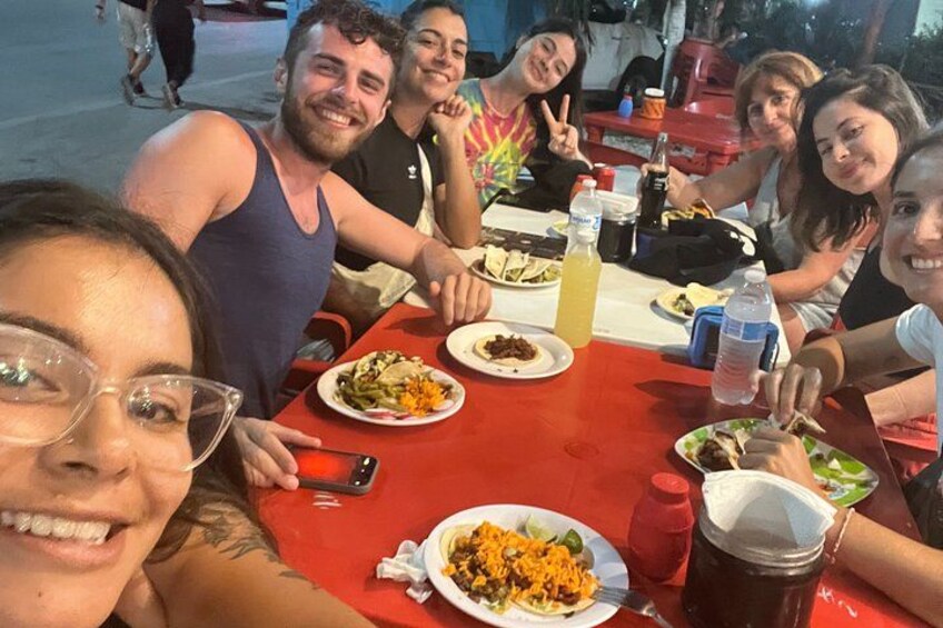 A group of people sitting at a red table with plates of food, drinks, and a smartphone on the table. 3