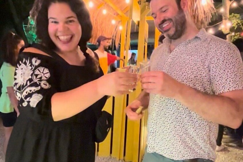 Two people toasting with drinks, a yellow gate, and a crowd in the background. 1
