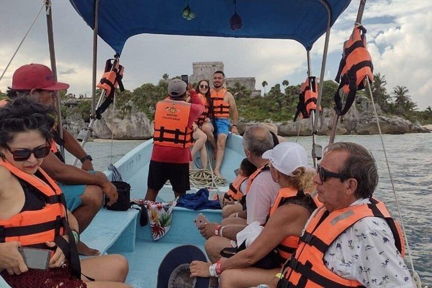 A group of people on a boat wearing life jackets, with a rocky shoreline and palm trees in the background. 5