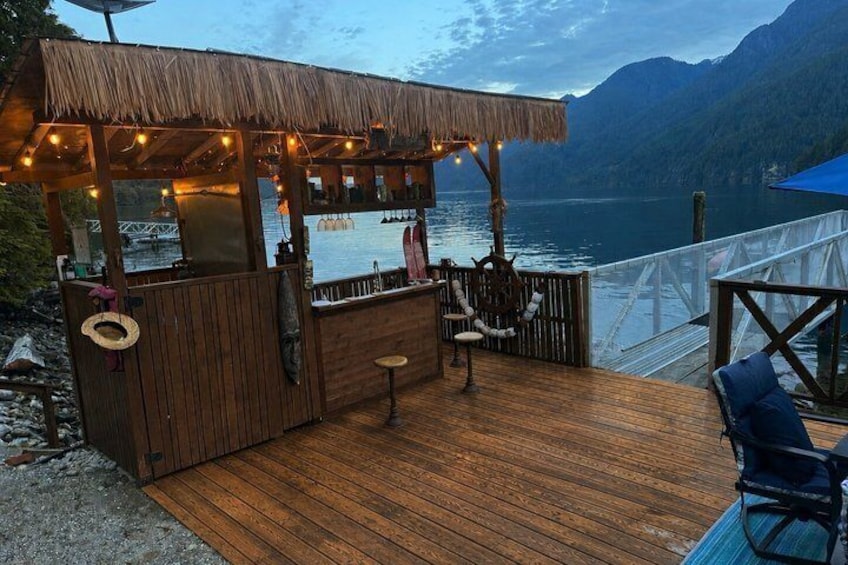 A wooden deck with a bar area, overlooking a calm lake and mountains. 3