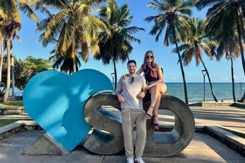 A couple posing with a large blue heart sculpture on a beach with palm trees and the ocean in the background. 1
