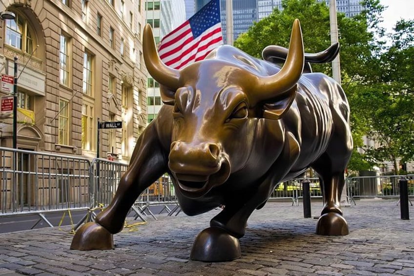 A bronze bull statue with an American flag, located on a cobblestone street with buildings in the background. 1