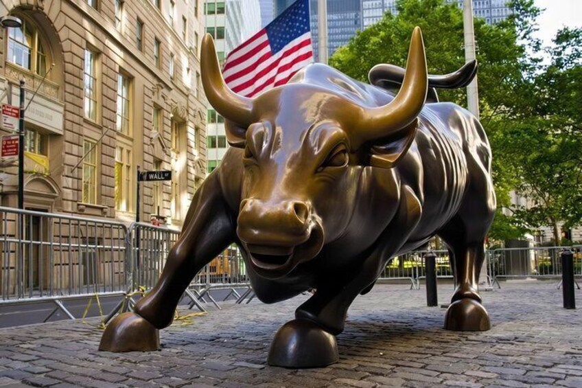 A bronze bull statue with an American flag, set on a cobblestone street with buildings in the background. 1