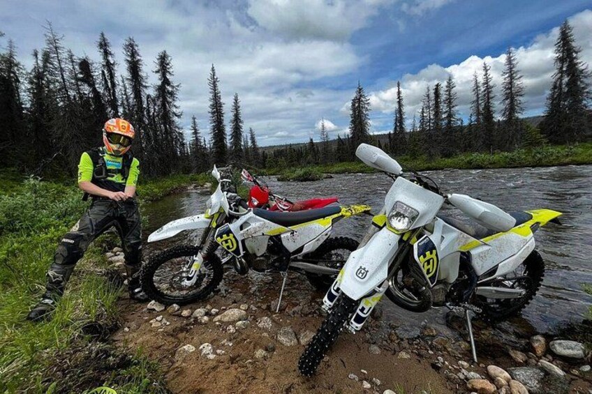 A person standing next to a white and yellow dirt bike by a river with trees in the background. 2