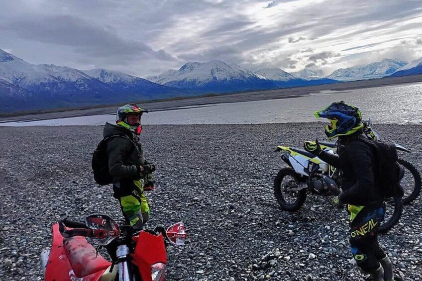 Two motorcyclists with helmets and backpacks on a rocky terrain with a lake and mountains in the background. 1