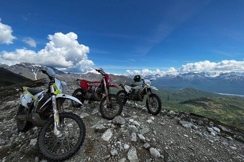 Three dirt bikes parked on a rocky mountain path with a scenic view of mountains and a lake. 4