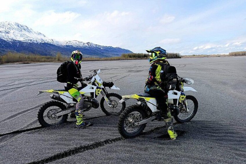Two people on motorcycles on a paved surface with mountains in the background. 3