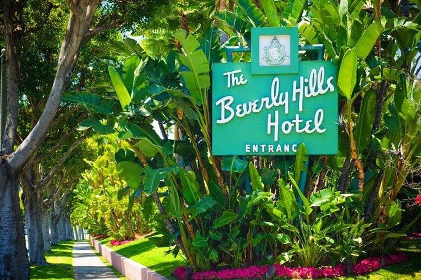 A sign for The Beverly Hills Hotel entrance surrounded by lush greenery and a pathway. 3