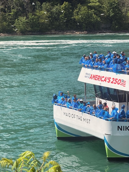 Vene, jossa on ihmisiä päällään siniset sadetakit, ja veneen kylkeen on maalattu tekstit »MAID OF THE MIST» ja »AMERICA'S 250th». Vene on vesistöllä. 2