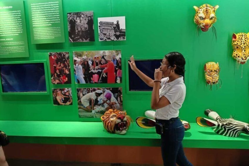 A person taking a photo in a museum with green walls, various masks, and informational displays. 3