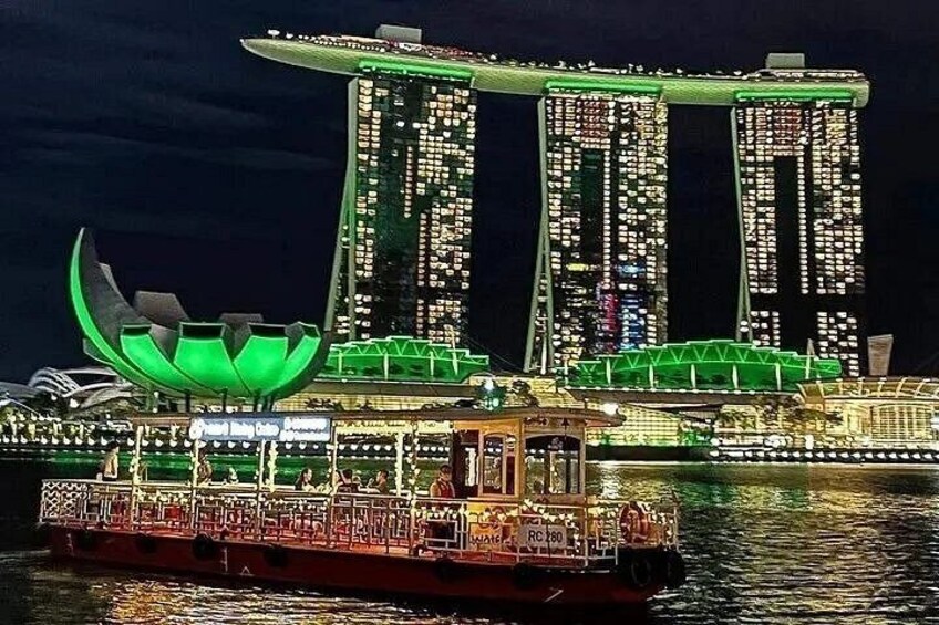En båt utsmyckad med upplysta gröna och vita lampor, en stor, grön lotusbladsformad konstruktion och Marina Bay Sands-hotellet med sina tre torn. 1