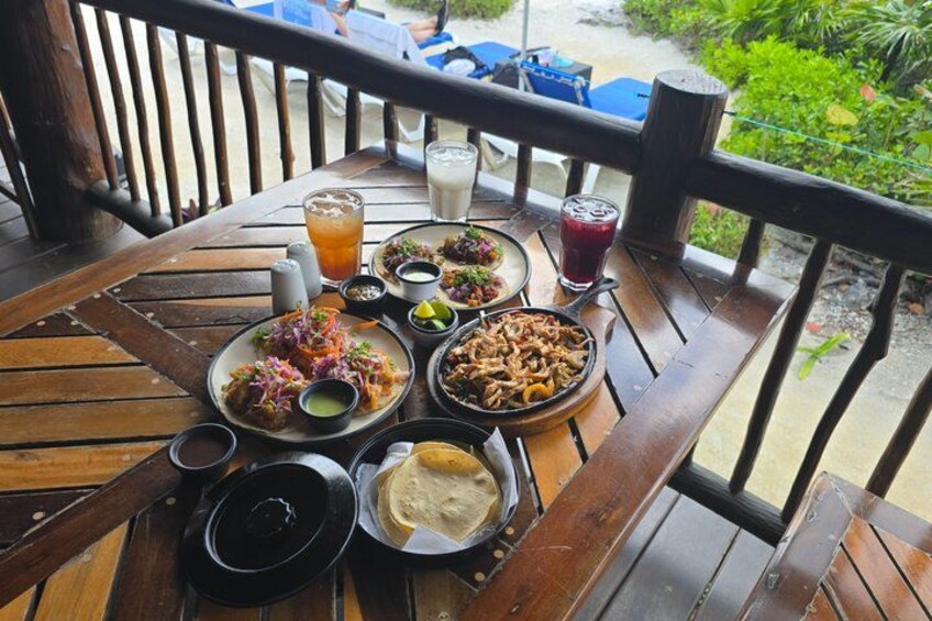 A wooden table with various dishes and drinks, surrounded by a wooden railing and greenery. 2