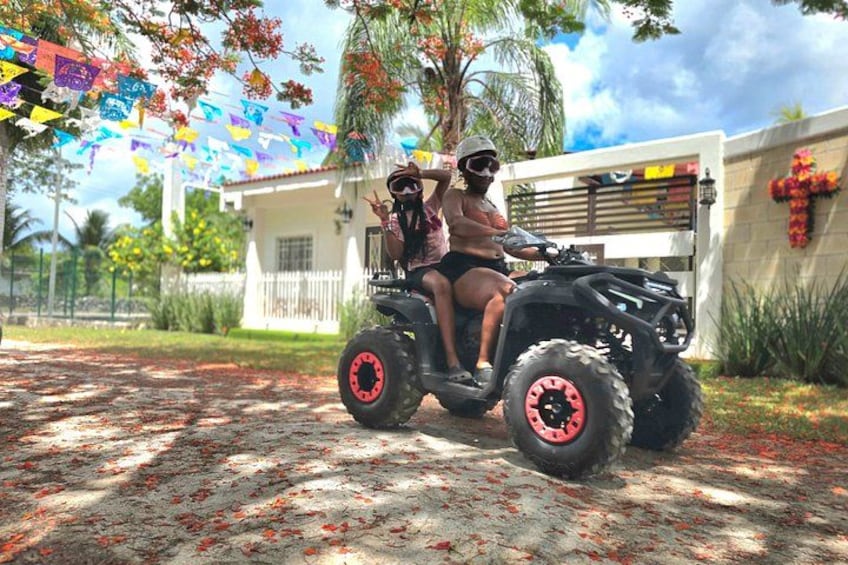 Two people on a quad bike in front of a house with a white fence and a red flower wall. 2