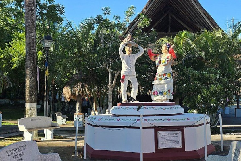 A statue of a man and woman on a pedestal with a plaque, surrounded by palm trees and a building with a thatched roof. 5
