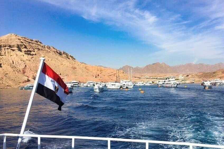 A flag on a boat, calm water, boats, and a mountainous landscape. 3