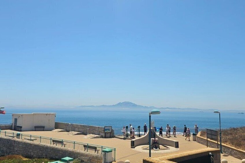 A coastal promenade with people walking, a clear sky, and a distant mountain. 2