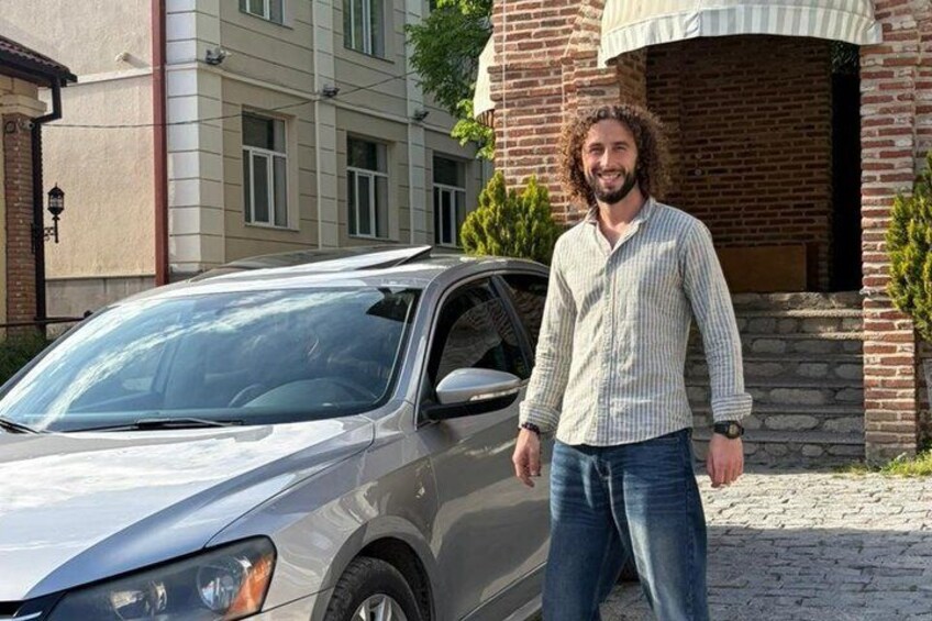 A man standing next to a silver car in front of a building with a brick facade and a window. 3