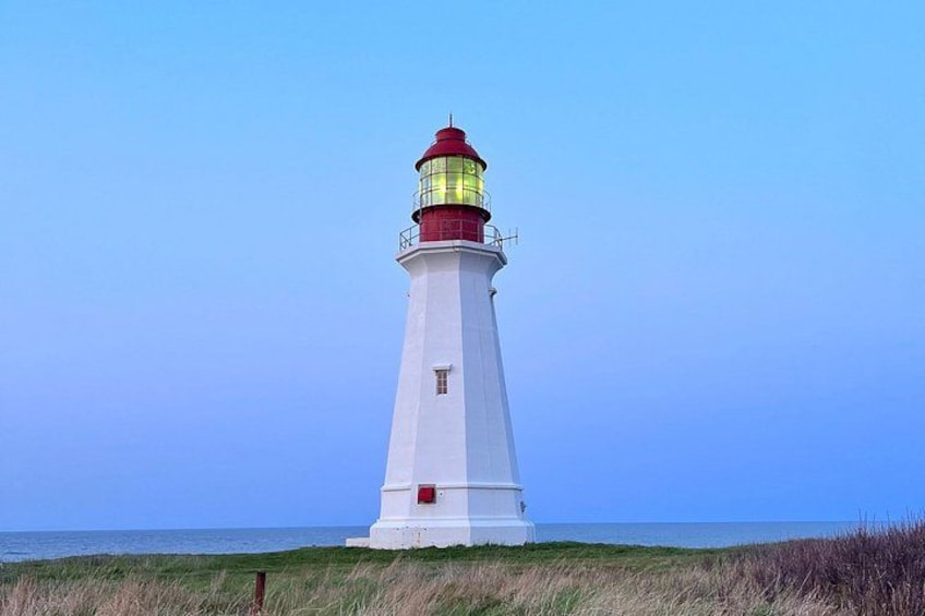 A white lighthouse with a red top and yellow light at the top, situated on a grassy beach with the ocean in the background. 4