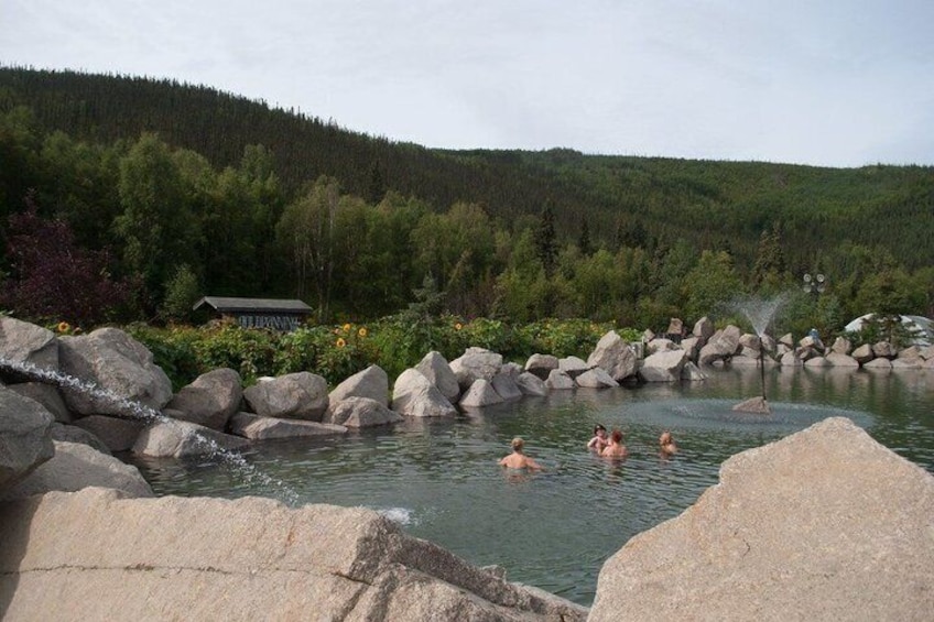 A natural hot spring with people swimming, surrounded by rocks and forested hills. 1