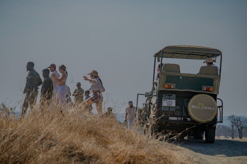 A group of people standing on a dirt path with a safari vehicle labeled 'HOLA AFRICA' in the background. 2