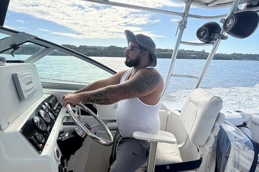 A person steering a boat on a body of water with a shoreline in the background. 2