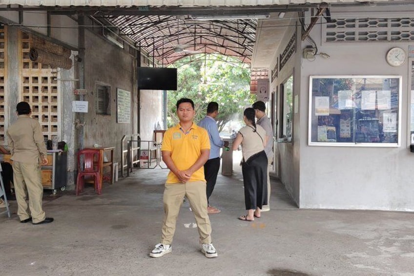 A man in a yellow shirt standing in a covered area with a clock on the wall, a red chair, and a bulletin board with papers. 1
